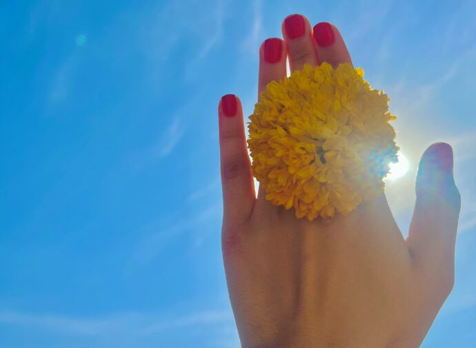 青空に花を挟んだ手を伸ばす赤ネイルをした手