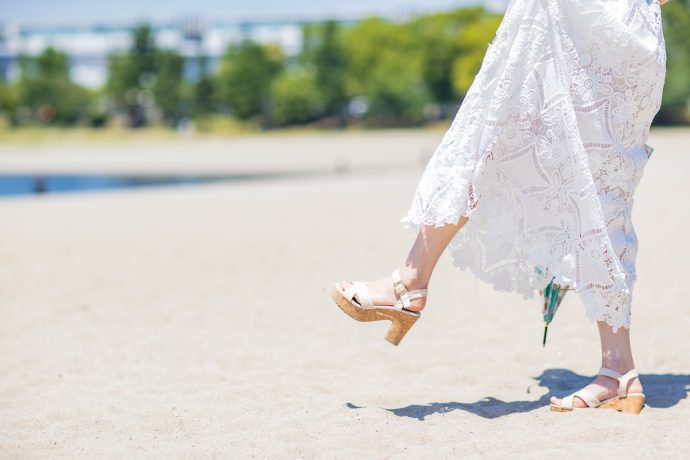 サンダルとロングスカートで晴れた日の砂浜を歩く女性の写真